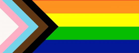 This image is of the updated Pride flag. The left side has white, pink, blue, brown, and black lines (in that order) creating a sideways "V" (like a greater than symbol) with the red, orange, yellow, green, blue, and purple horizontal stripes to the right.